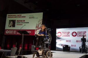 maayan smiles on stage at the startup canada day on the hill event. behind her is a large screen that reads "Announcement: Startup Jerusalem Competition Canada" in red