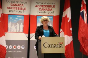 The Hon. Carla Qualtrough stands at a podium branded with the Government of Canada logo in front of Canadian flags at Abilities Centre.