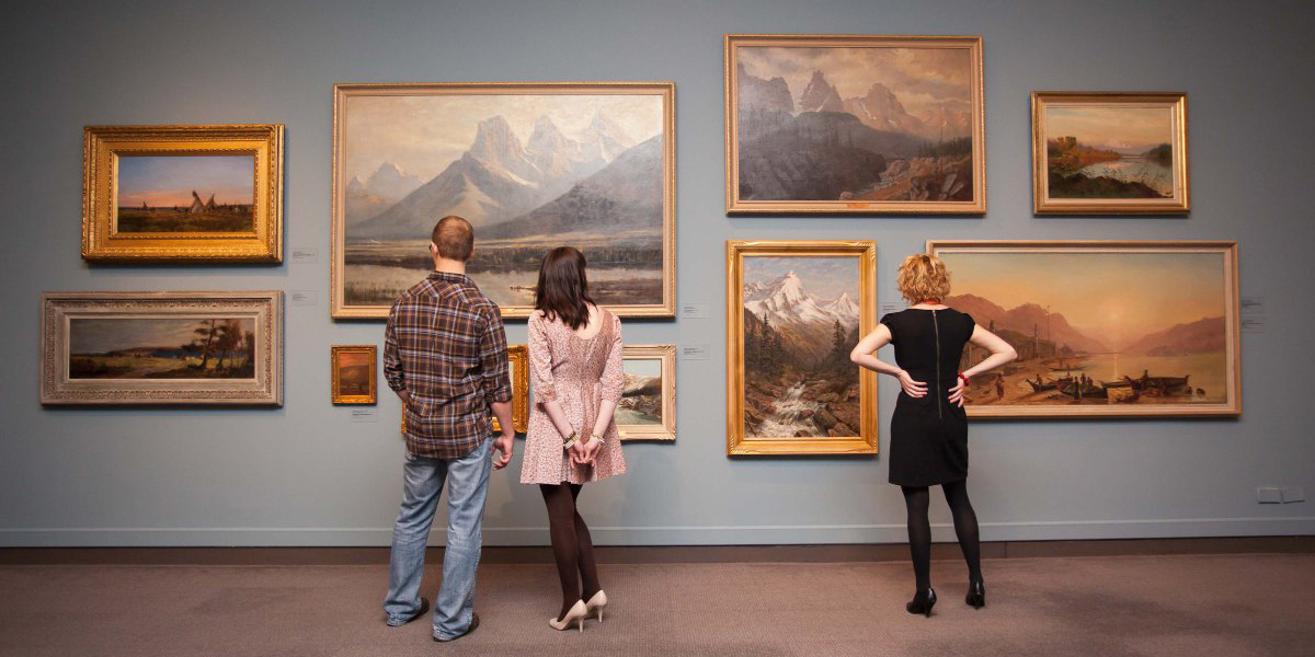 three people stand looking at a wall of landscape paintings in a museum