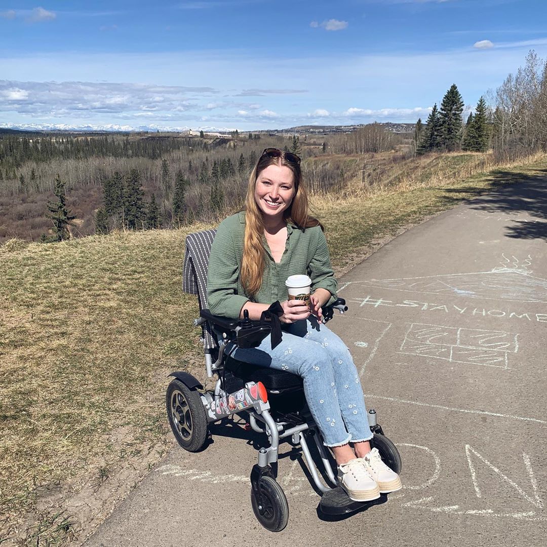 Kelcie sitting in a wheelchair outdoors, prairies in the background. She is looking at the camera, smiling and with a cup of coffee in hand. 