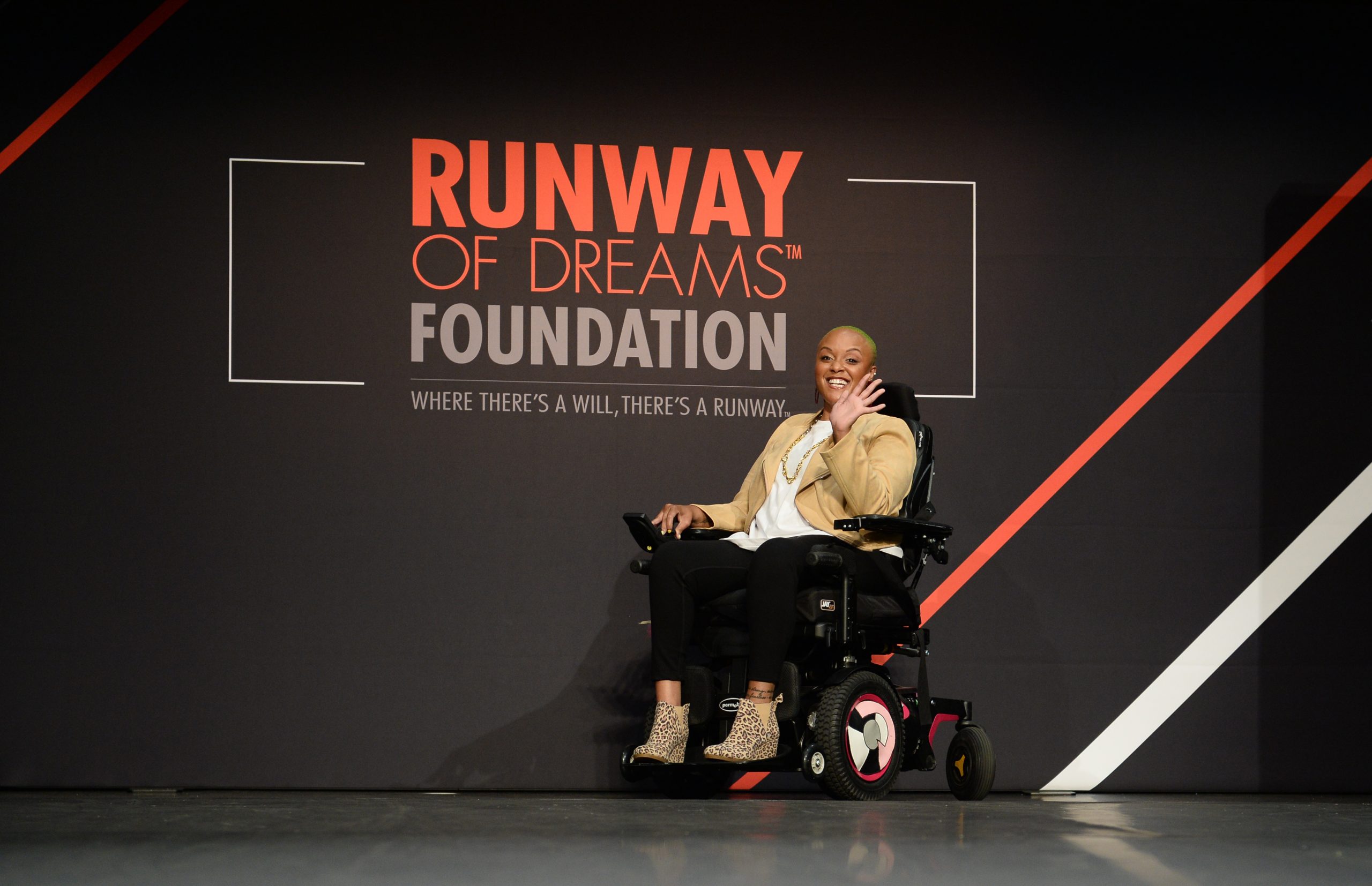 Keisha in a wheelchair in the middle of a fashion runway. She is waving and smiling at the camera. There is a black backdrop behind her that says “Runway of Dreams Foundation: Where there’s a will, there’s a runway.”