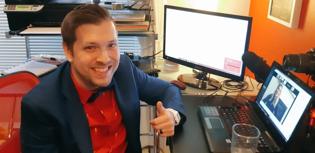 Marco dressed up formally with a suit and a bowtie. He is looking at the camera, smiling and giving a thumbs up. He is at home, in his office and surrounded by computer screens.