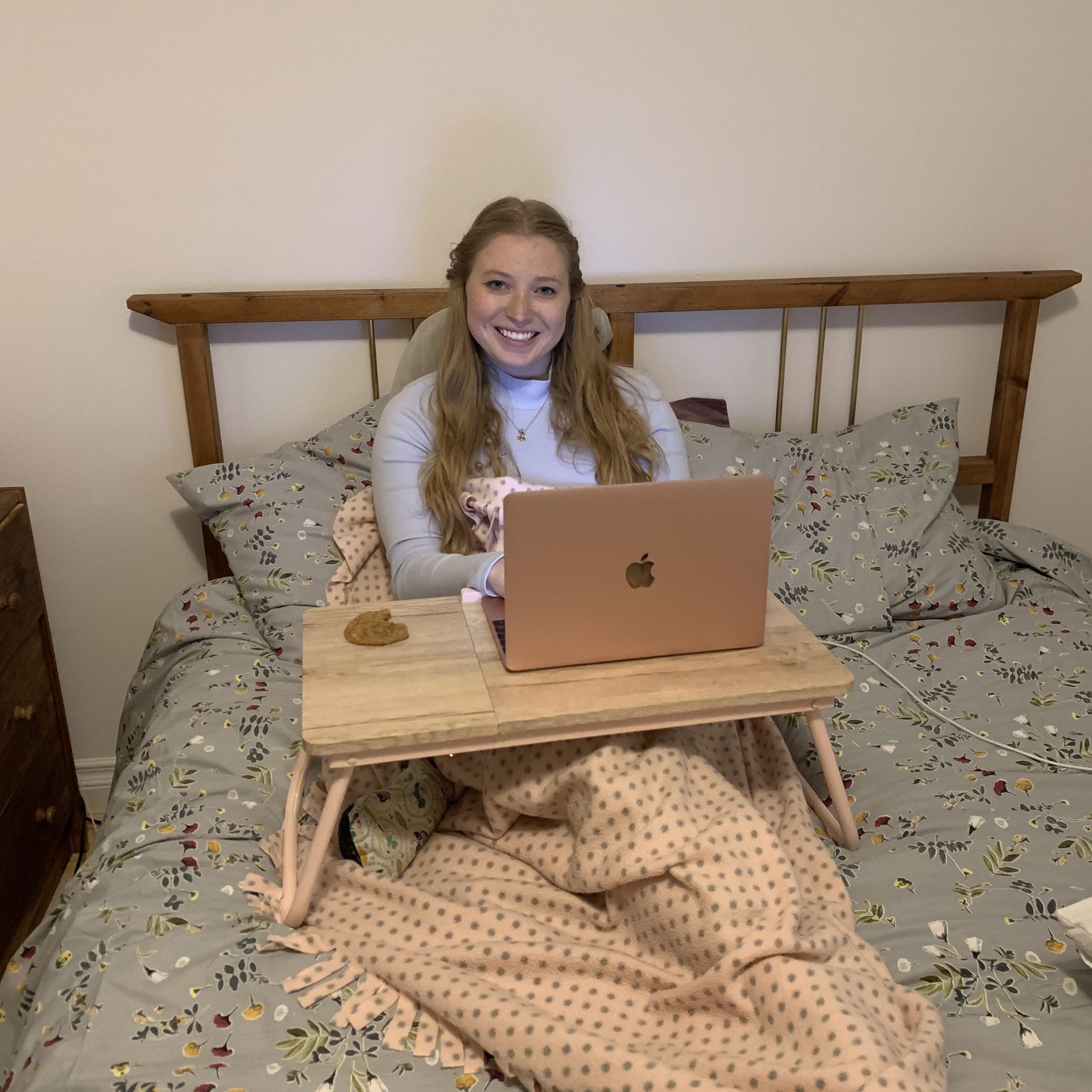 Kelcie sitting up on her bed. She looks comfortable and wrapped in blankets. There is a bed tray propped in front of her with her laptop on it. The laptop is open and you can see the light from the screen hitting her face. She is smiling and looking directly at the camera.