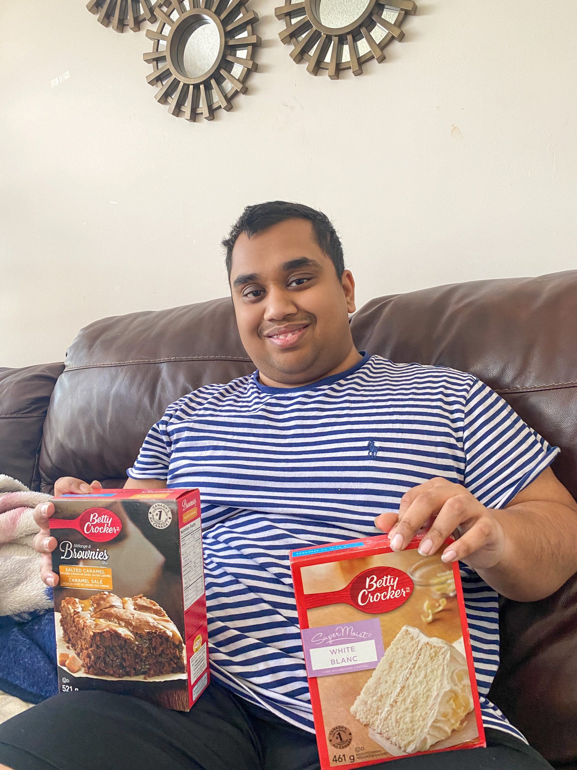 Ridwan sitting on a couch and smiling at the camera. He is holding boxes of Betty Crocker baking mixes. One hand has a brownie mix and the other a cake mix. 