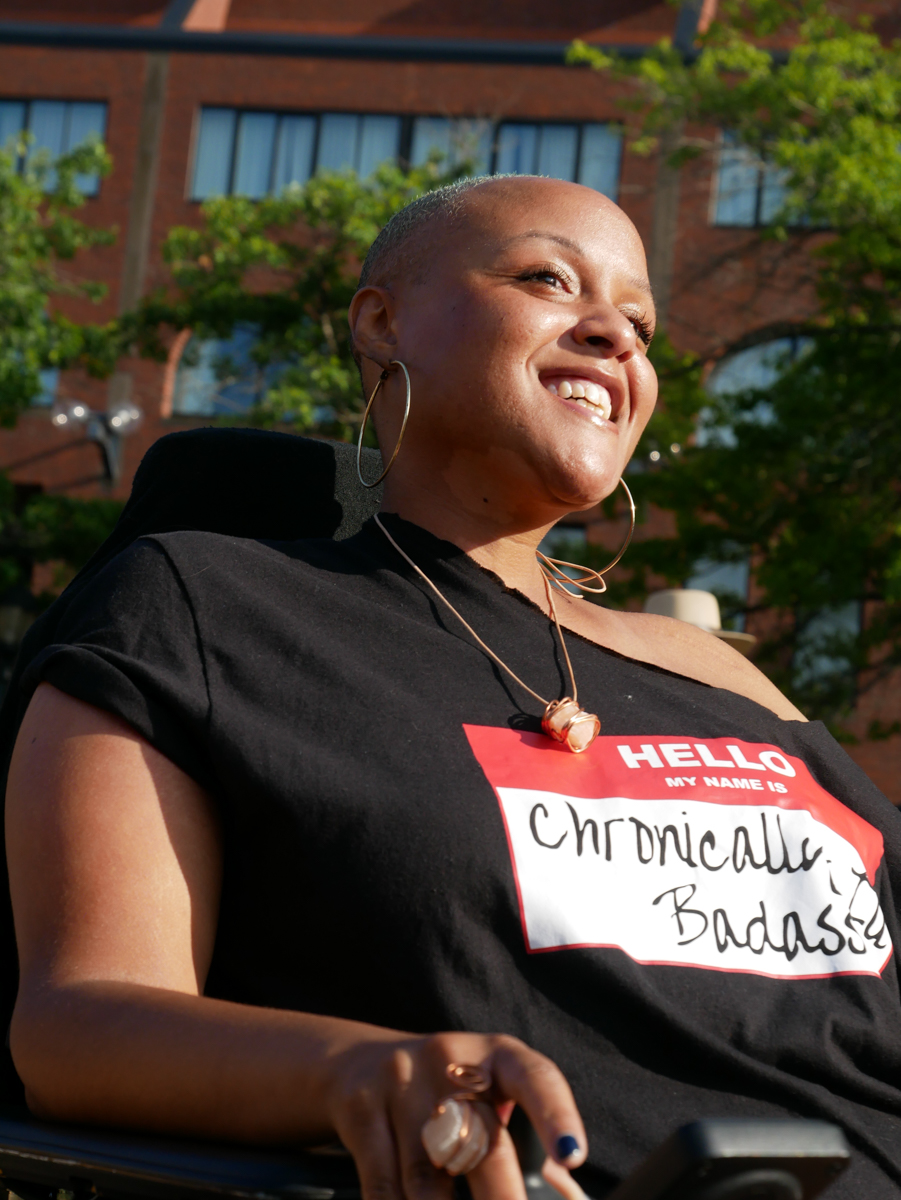 Portrait of Keisha from the waist up. Light hits her face and she is smiling. She has hoop earrings and a necklace. She is wearing a black off-shoulder top that says “Hello, My name is: Chronically Badass”. 