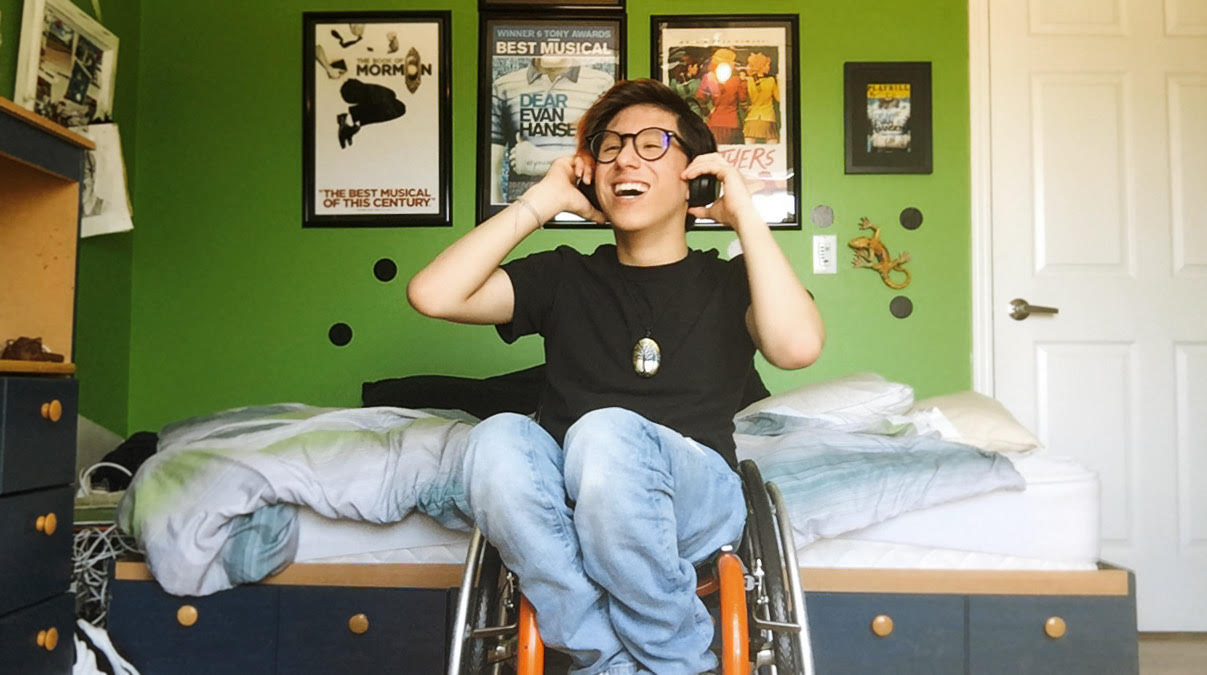 Portrait of Tai Young sitting in his wheelchair inside his bedroom. He has headphones on and has his hands up to touch it. He is smiling.