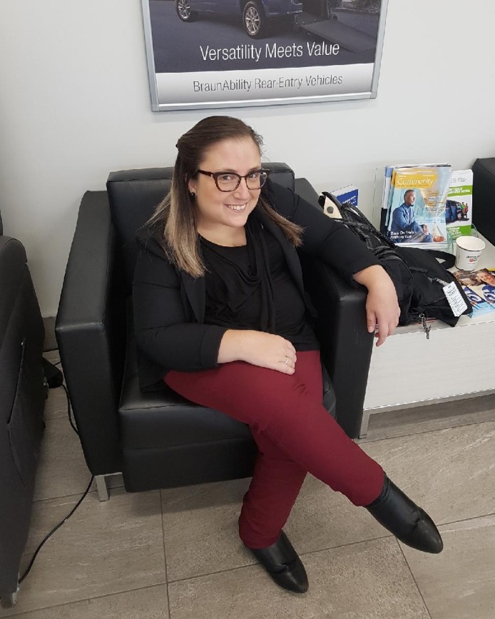 Gabby sitting in a low black lounge chair with her legs crossed. She is looking at the camera and smiling. She has her hair pinned to the back, she is wearing glasses, a black cardigan, maroon pants and black boots.