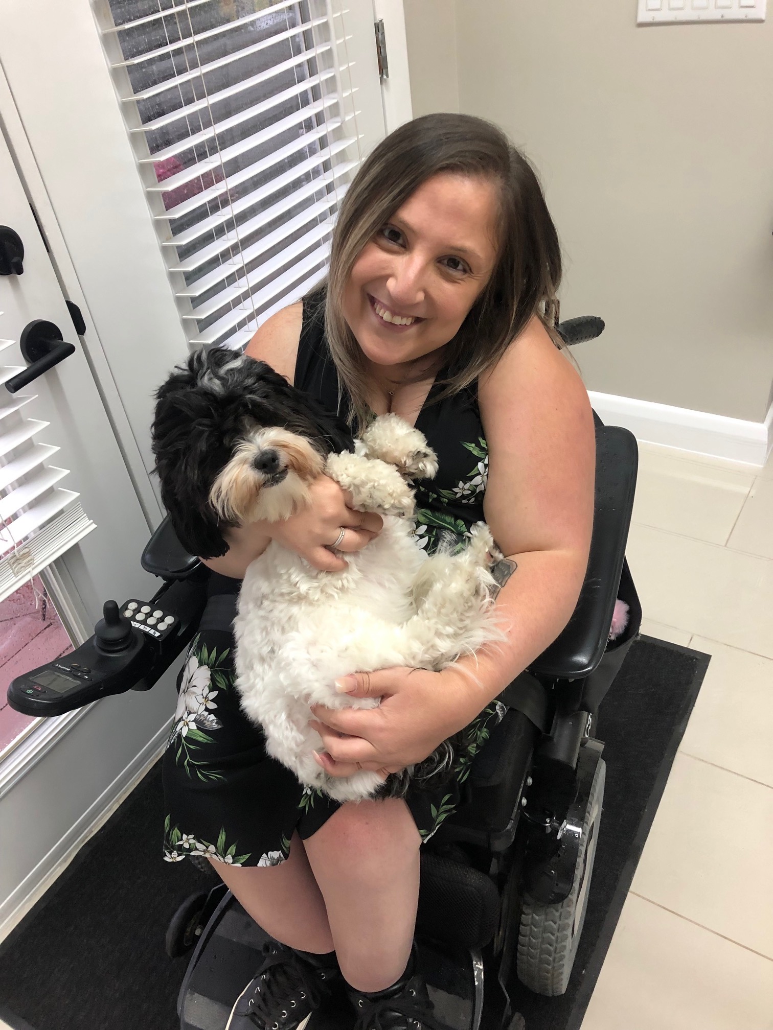 Gabby in her wheelchair. She is looking at the camera with a sweet smile on her face while she is holding a dog on her lap.