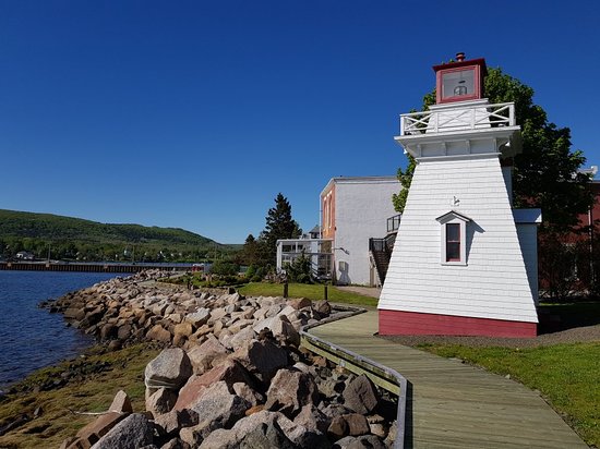 A wooden boardwalk trail that leads to a white and red lighthouse, named the Annapolis Royal lighthouse. The boardwalk winds around a bright blue body of water with a rocky beach. 
