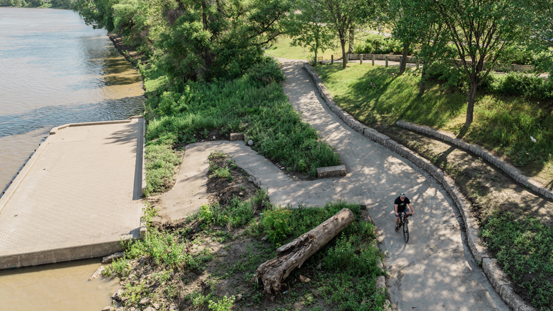 A bird's eye view of someone biking along a smooth paved path, which has plenty of greenery surrounding it. There is a small path that branches out from the main pathway and leads to a concrete lookout area on the water.