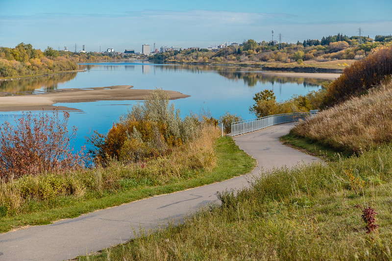 A smooth, paved pathway that loops along a calm body of water. There is plenty of greenery surrounding the path, and off in the distance you can see the skyline from the city. 