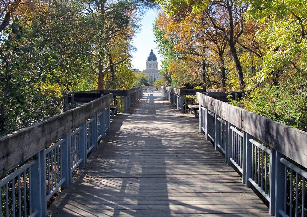 A wooden planked pathway with luscious green, orange and yellow trees surrounding it on both sides. The pathway leads to the Saskatchewan Legislative Building, which you can see off in the distance. 