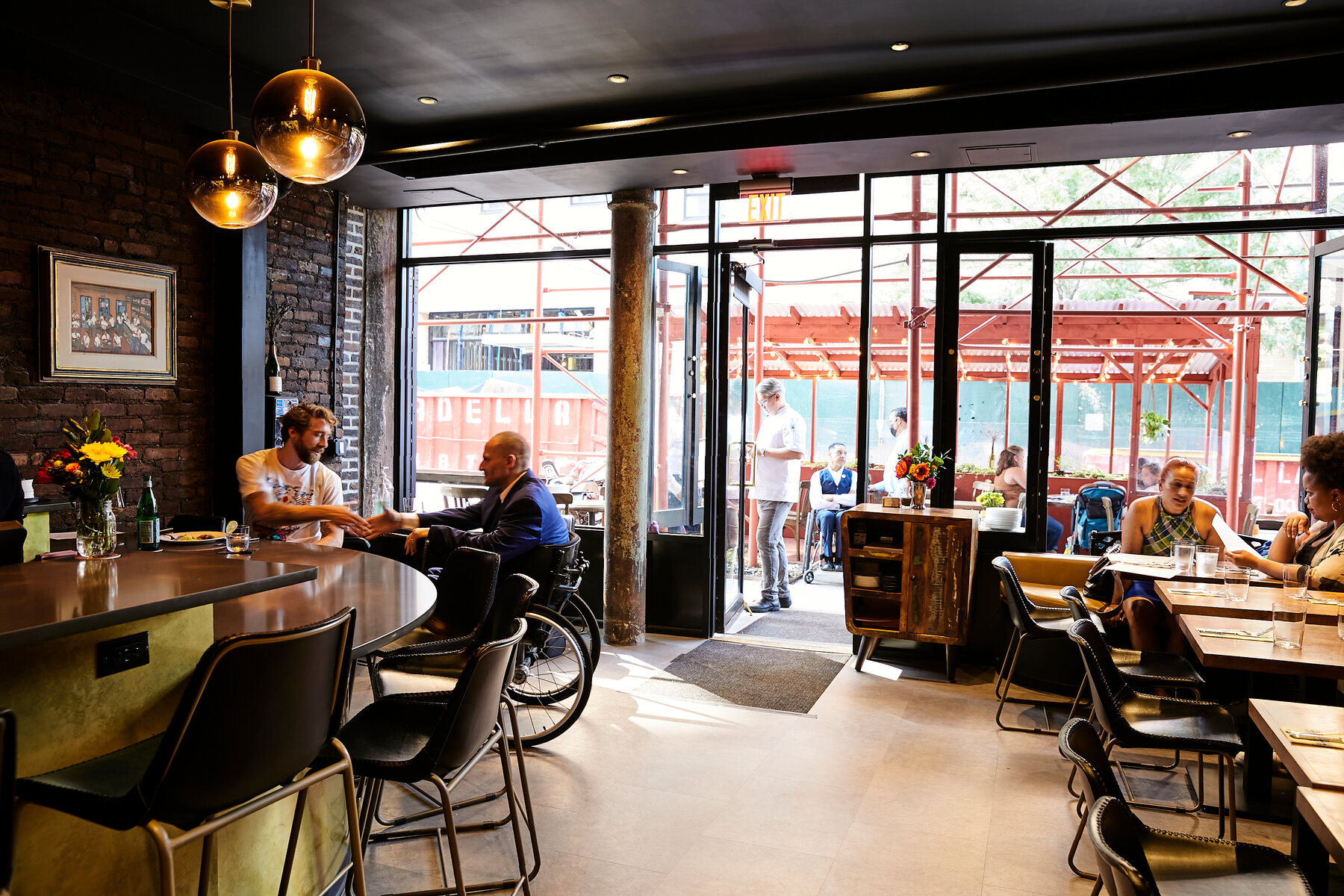 This photo shows the inside Contento restaurant. It is an open concept design with tables spaced out along the right hand side, and the bar against the left hand side. There is a clear pathway down the middle. The owner of the restaurant, Yannick Benjamin, is shaking hands with a guest who is seated at the lower half of the bar. Outside the doors, there is a patio with numerous diners sitting out there.