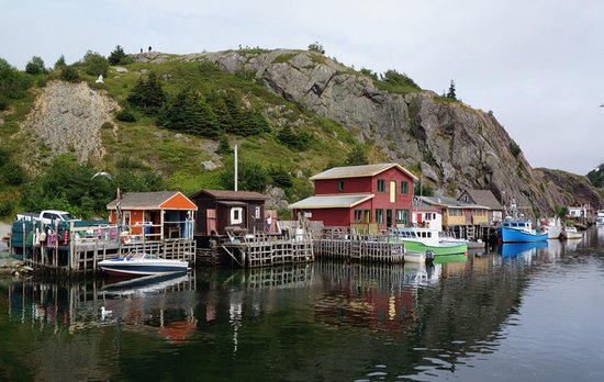 The shoreline of Quidi Vidi in St. John’s. There are several brightly coloured boat houses along the shore, with wooden docks and fishing boats in front of each. There is a large rocky cliff in the background. 