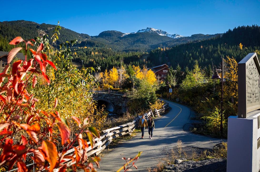 Two individuals walking on the Whistler Valley Trail. The trail is a smooth asphalt pathway, and you can see large mountains in the background. The photo appears to be taken in the Fall as the trees are various colours of green, red and yellow. 