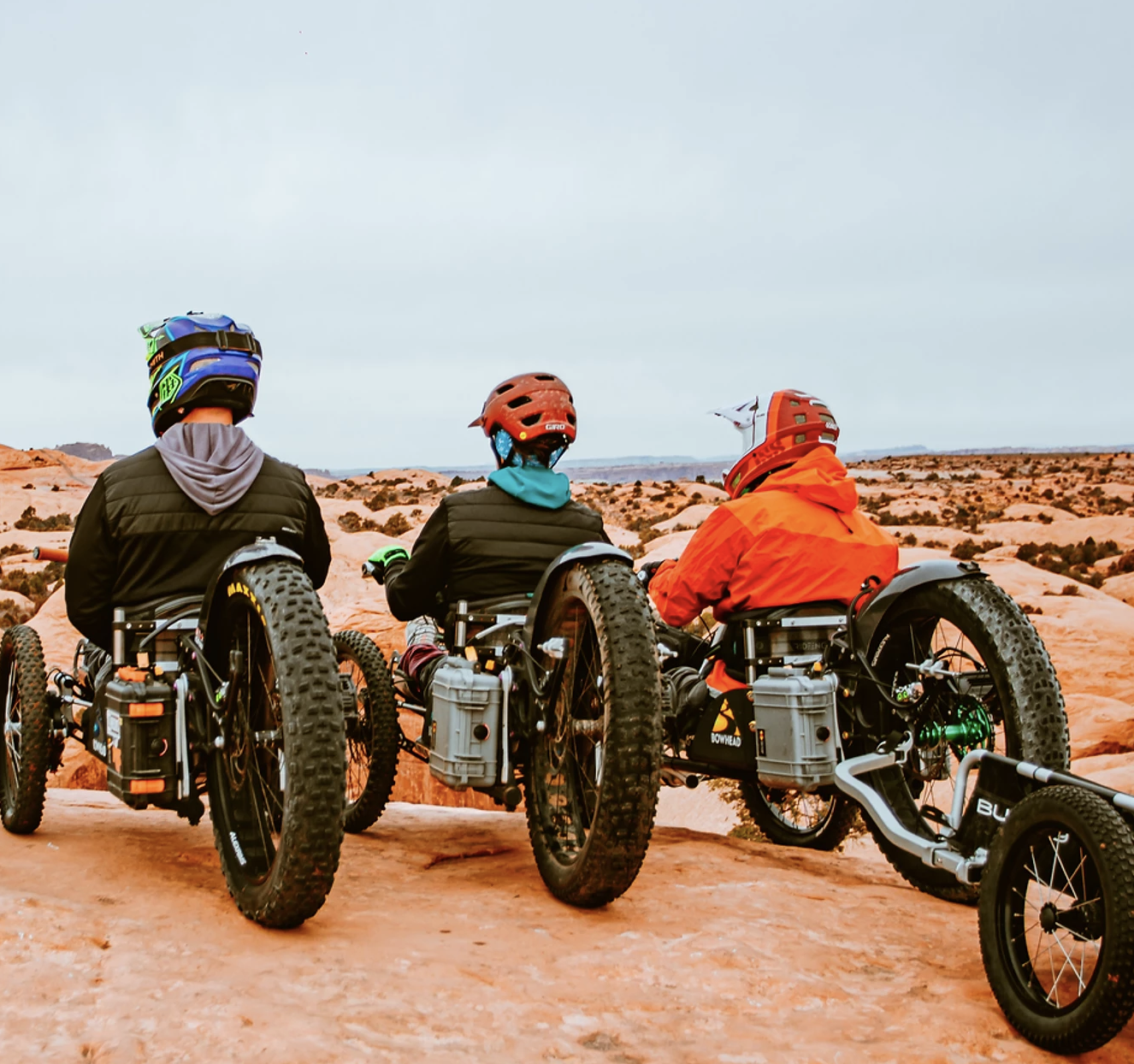 3 dudes on adaptive bikes sit on a sandy hill overlooking a view