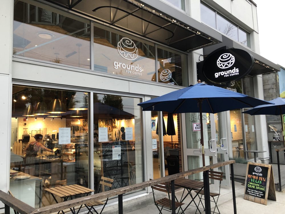 A photo of the outside of grounds for coffee. There is a patio area out front of the cafe which has two small tables and chairs. There is a handwritten chalk sign out front that says “try our new vegan chocolate chunk brownie”. Through the window, you can see the barista inside working at the service counter.