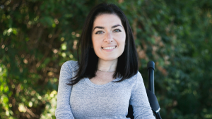image of maayan ziv smiling in her wheelchair. she has brown hair and sits comfortably in a park.