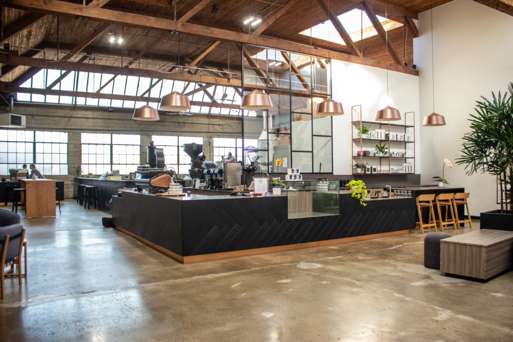 A photo of the inside of pallet coffee roasters. The space is very large and warehouse-like. The floors are polished concrete and the service bar is black with gold accents and gold chairs. The exposed ceilings are very high, showing the wood foundation. 