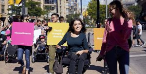 maayan smiles in a sunny street. she drives her power wheelchair while leading a team of interabled people behind her who carry colourful signs that read "there is strength in diversity"