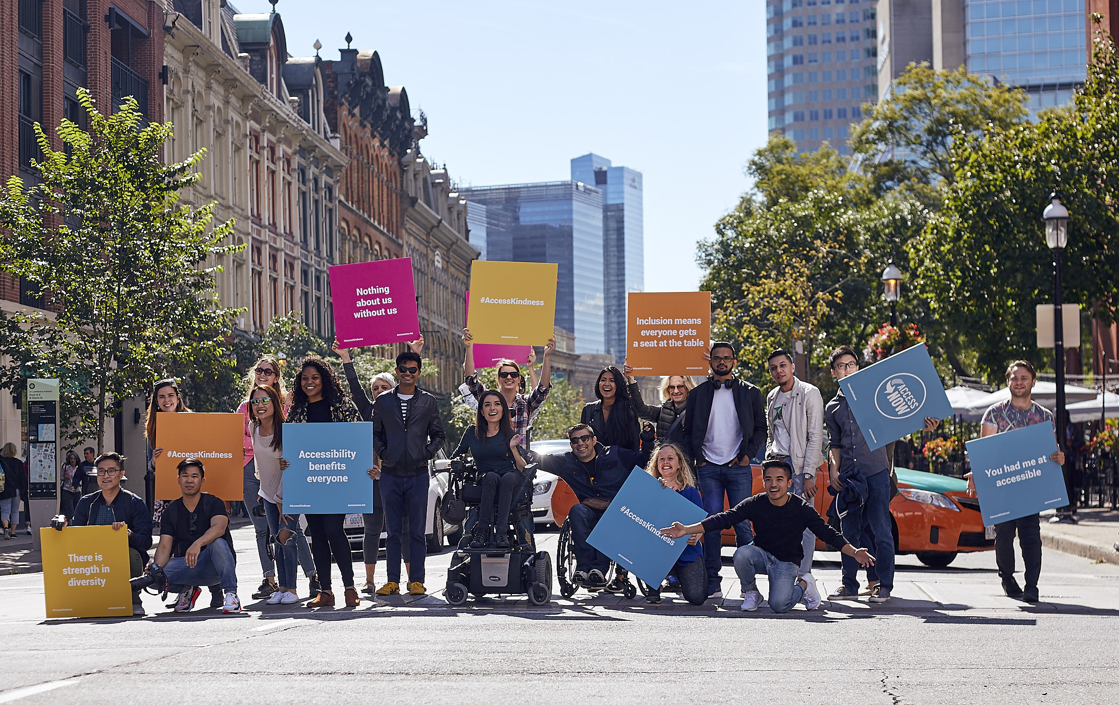 Group of people with disabilities and allies stopping traffic in downtown Toronto. They are holding up various signs that promote kindness.
