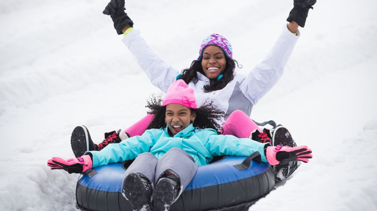 A mom and daughter sliding down a snowy hill in a snow tube. The child is sitting in front, in between her mom's legs. They are both laughing in excitement. 