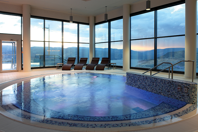 A large indoor hot tub at Sparkling Hill Resort. It is surrounded by floor to ceiling glass windows, and outside, you can see the silhouette of large mountains.