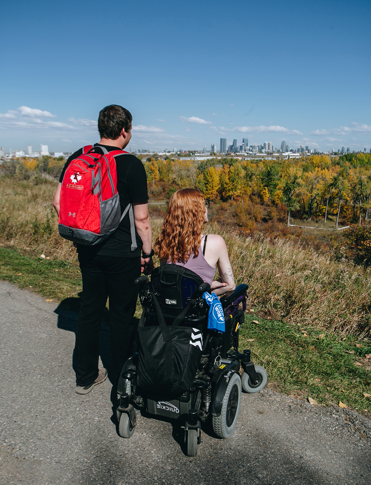 two people look out over a park at the city of calgary.  on a sunny day. one person is sitting on a power wheelchair.