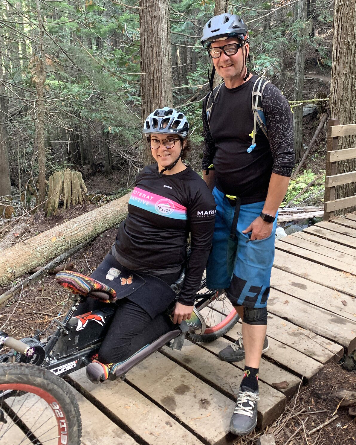 kim, left, smiles in her hand cycle, ormond, right, stands next to her on a boardwalk