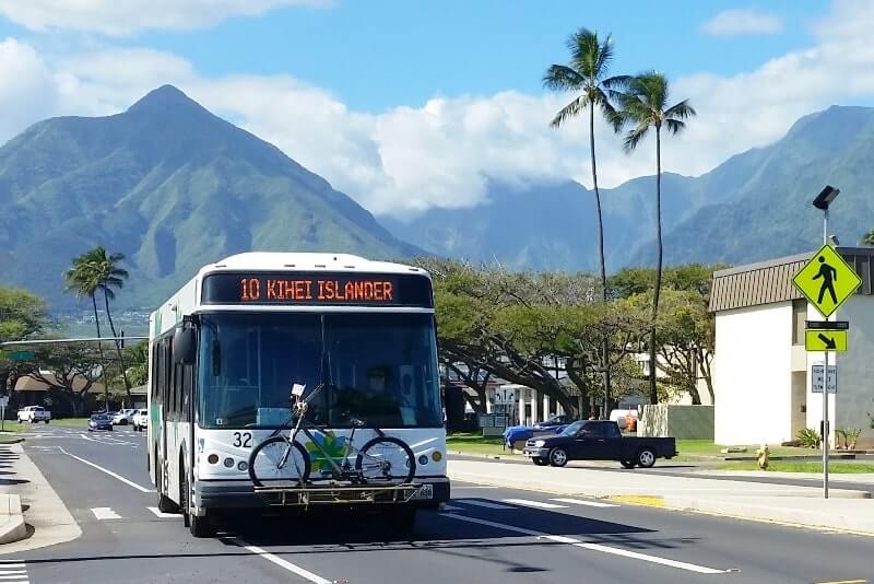 Maui public bus driving on a road in Maui. Behind the bus there is a scenic background full of palm trees and mountain peaks. 