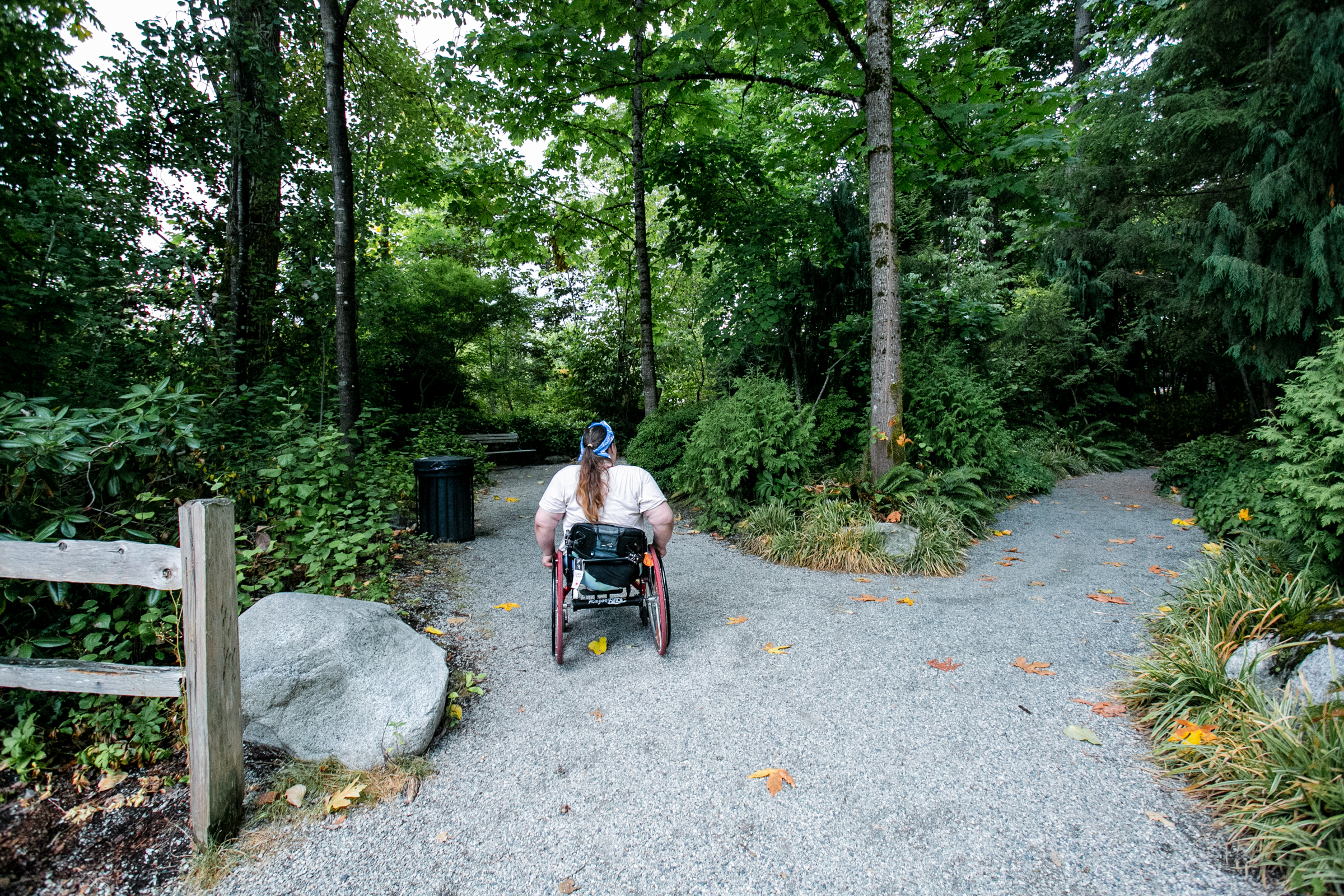 Two individuals walking on the Whistler Valley Trail. The trail is a smooth asphalt pathway, and you can see large mountains in the background. The photo appears to be taken in the Fall as the trees are various colours of green, red and yellow. 