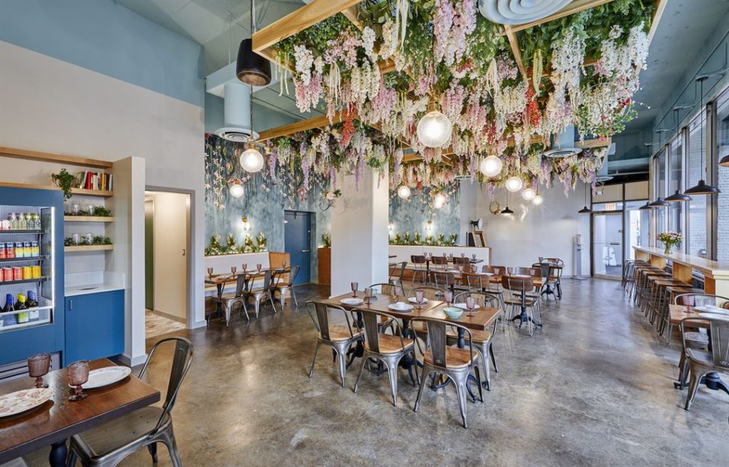 Beautiful interior of a restaurant. High ceilings, with floral decorations hanging from the ceiling. Lots of natural light and very spacious aisles between tables.