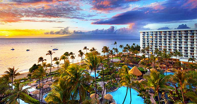 A scenic drone view of a hotels property. There are numerous pools, cabanas, and palm trees in sight. In the background you can see the Kaanapali Beach front, and the sky is filled with vibrant purple and blue hues with the sun setting in the horizon. 