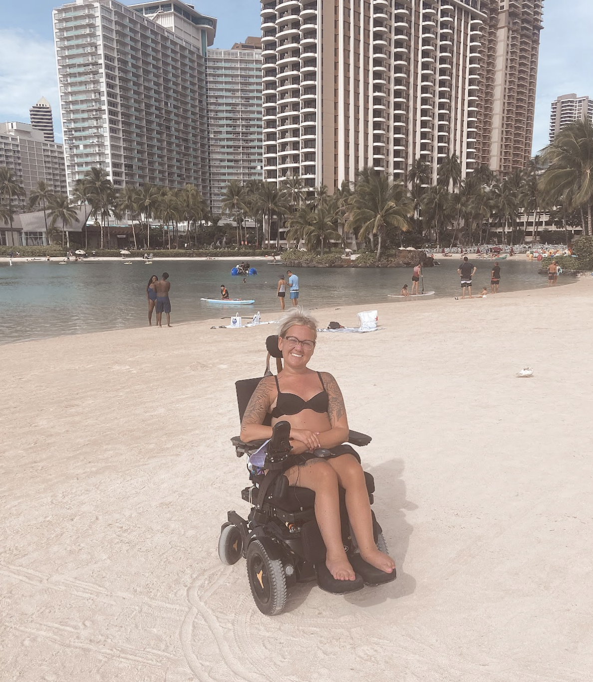 Shailynn sitting on her wheelchair. She is at the beach, wearing a black bikini and eye glasses. Behind her is a lagon and a high-terrace building. There are people in the lagoon.