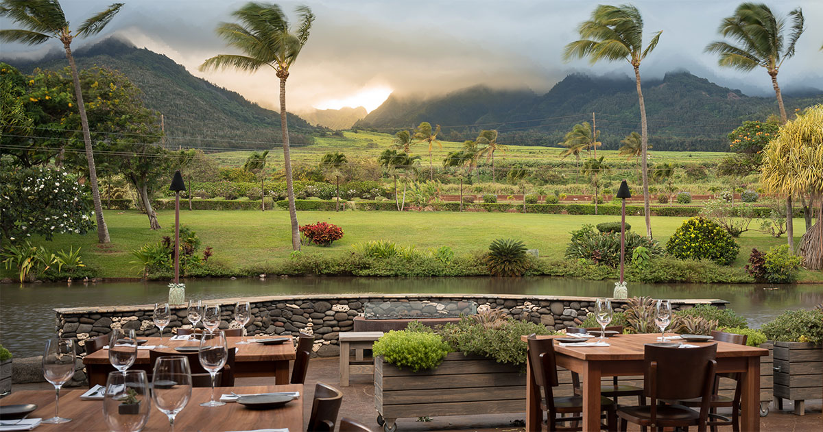 Tables, chairs, and place settings set up on the patio of Cafe O’Lie at The Mill House (located on the property of the Maui Tropical Plantation). The tables overlook a very scenic view of the West Maui mountains, as well as the pond and greenery located on the property.   