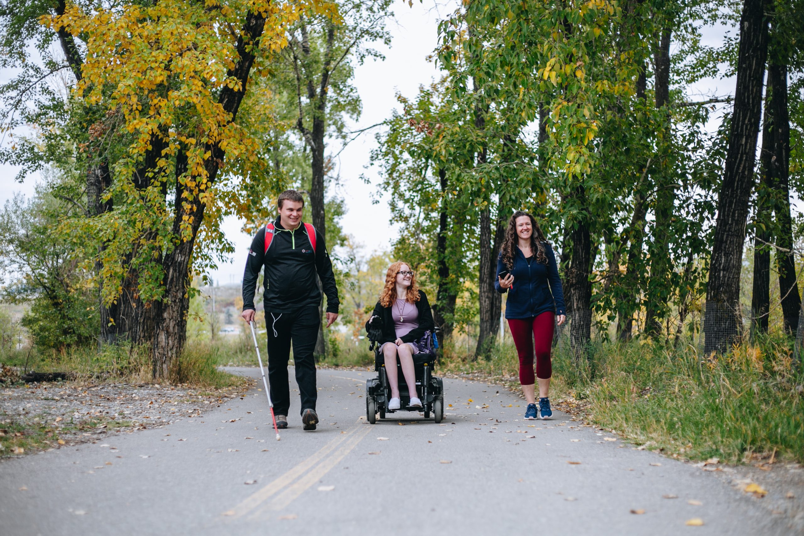 Accessibility mappers, a young blind man walking with a cane and a young girl sitting on a wheelchair, with their trail guide. They are walking on a paved path in a park on a beautiful autumn day.