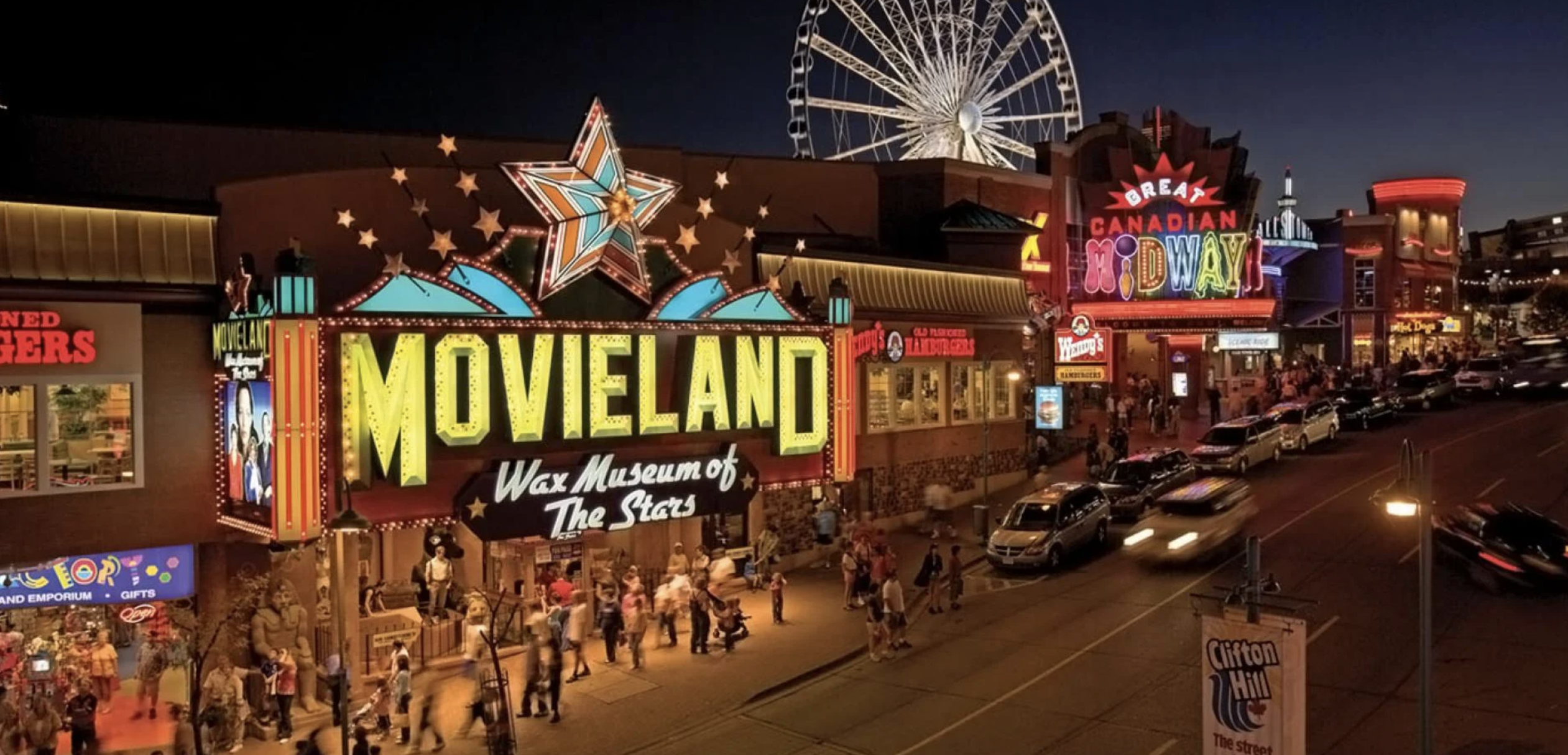 Clifton Hill at night. The street is filled with tourists and vibrant with life. Buildings with neon lights and signs. Ferris wheel lit up at the background.