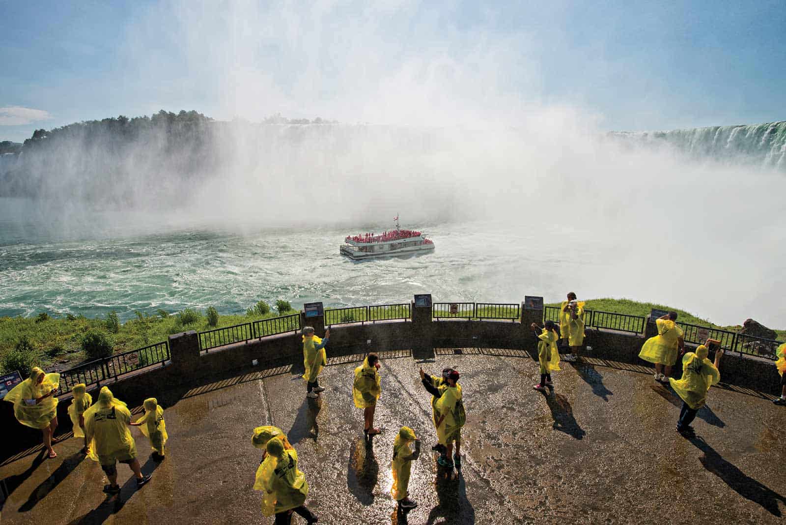 View of the falls. There is mist coming out and a boat in the middle of the lake. There is an observation deck with people wearing yellow ponchos, a lot of them taking pictures
