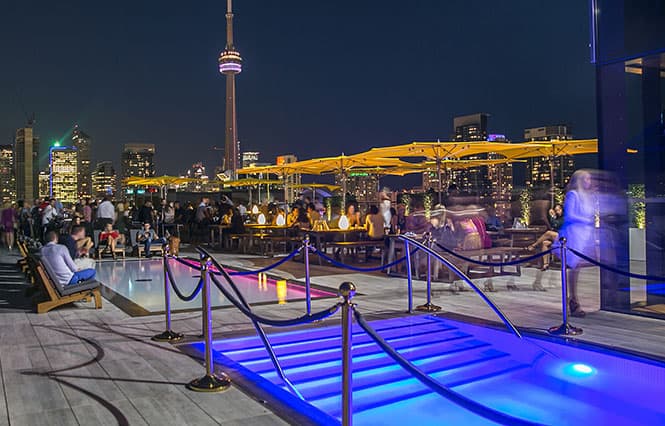 busy rooftop bar at night. there is a pool with lights. CN tower lit up at the back.