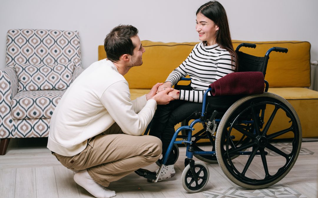 Woman sitting in a wheelchair, man crouching down holding her hand. They are both smiling.