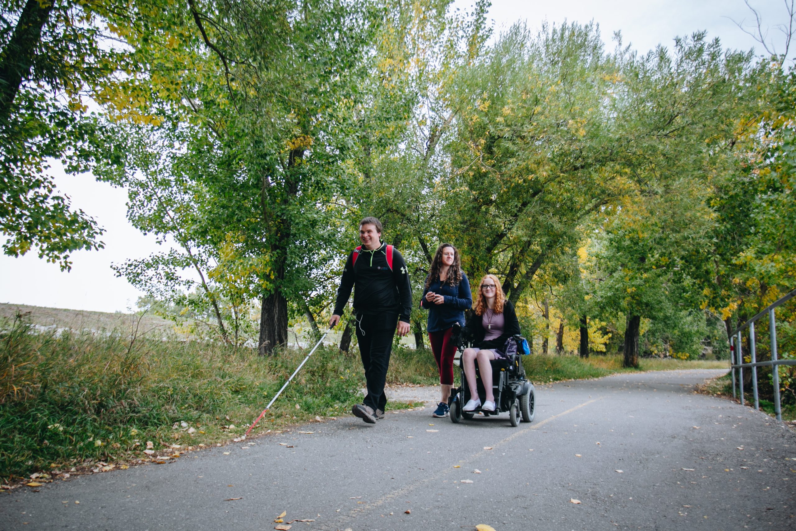 Woman in a power wheelchair and a man using a whitecane, stroll along a paved path in a forested area