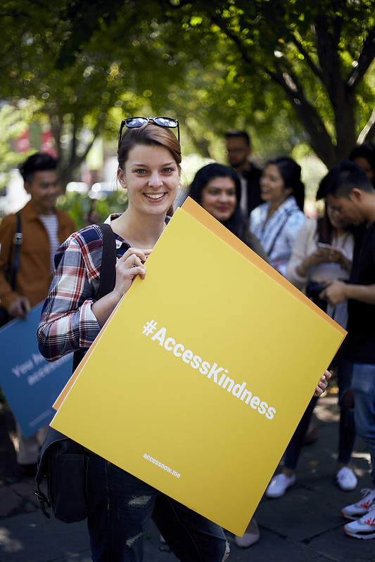 photo of young woman smiling as she holds up a yellow sign that reads "Access Kindness" in bold 