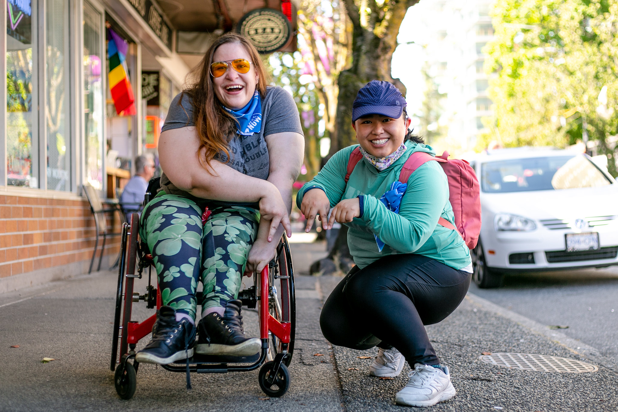 deux femmes, dont l'une est assise sur un fauteuil roulant, sourient en posant sur le trottoir pour l'appareil photo