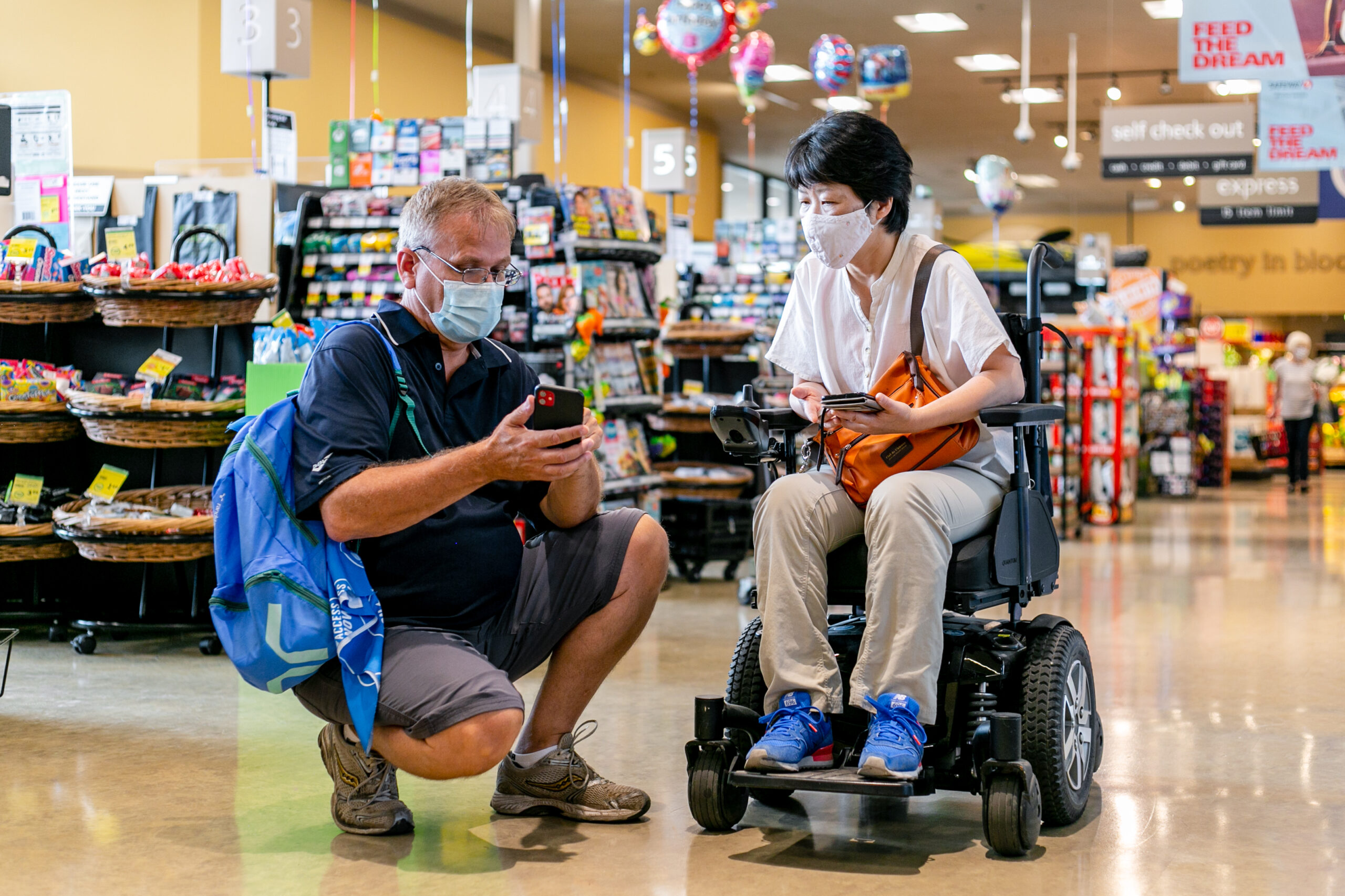 an asian woman sitting in a wheelchair and a white man both look at a mobile phone while inside a grocery store