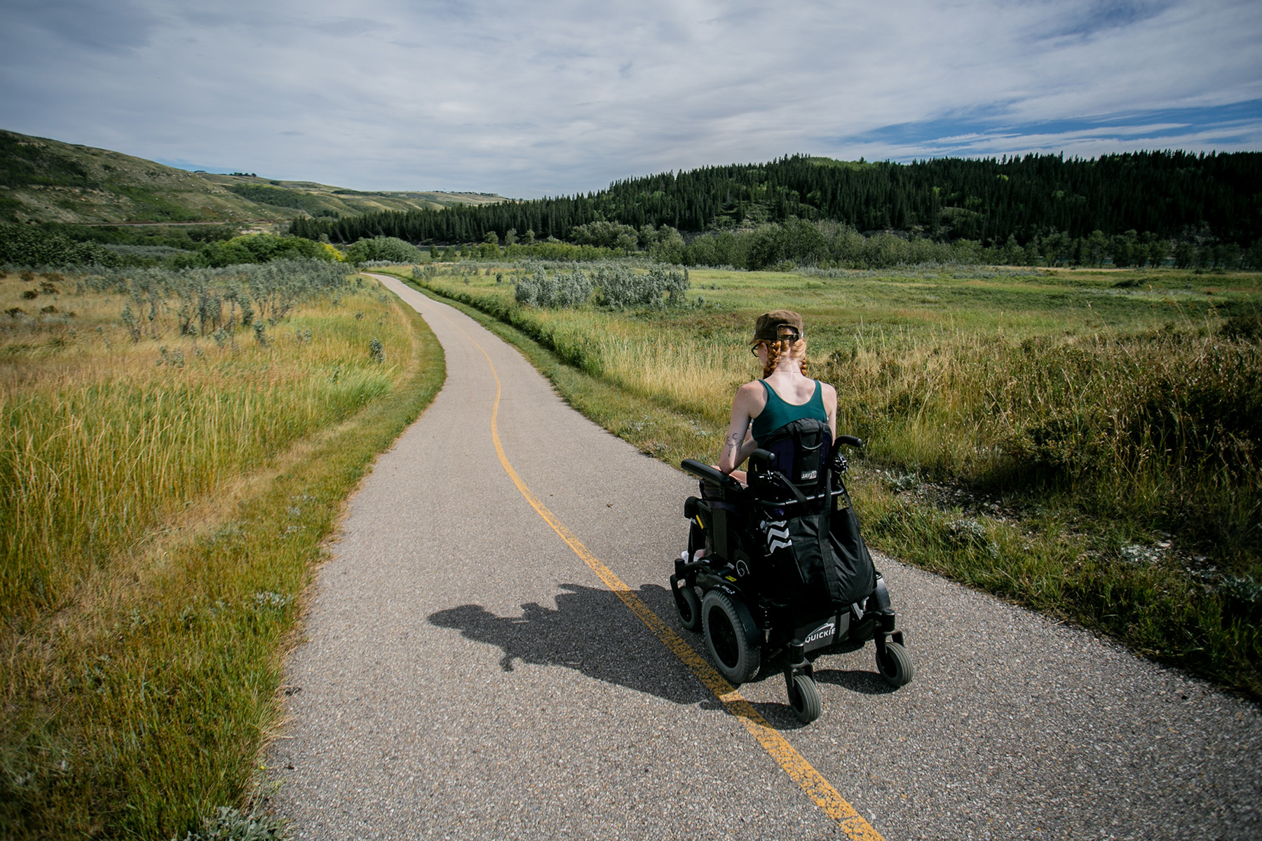 a woman in a powerwheelchair is seen from the back wheeling on an open paved trail within a green field