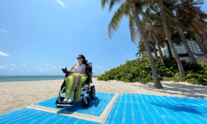 woman in wheelchair poses on beach mat. behind her is a sandy beach and ocean