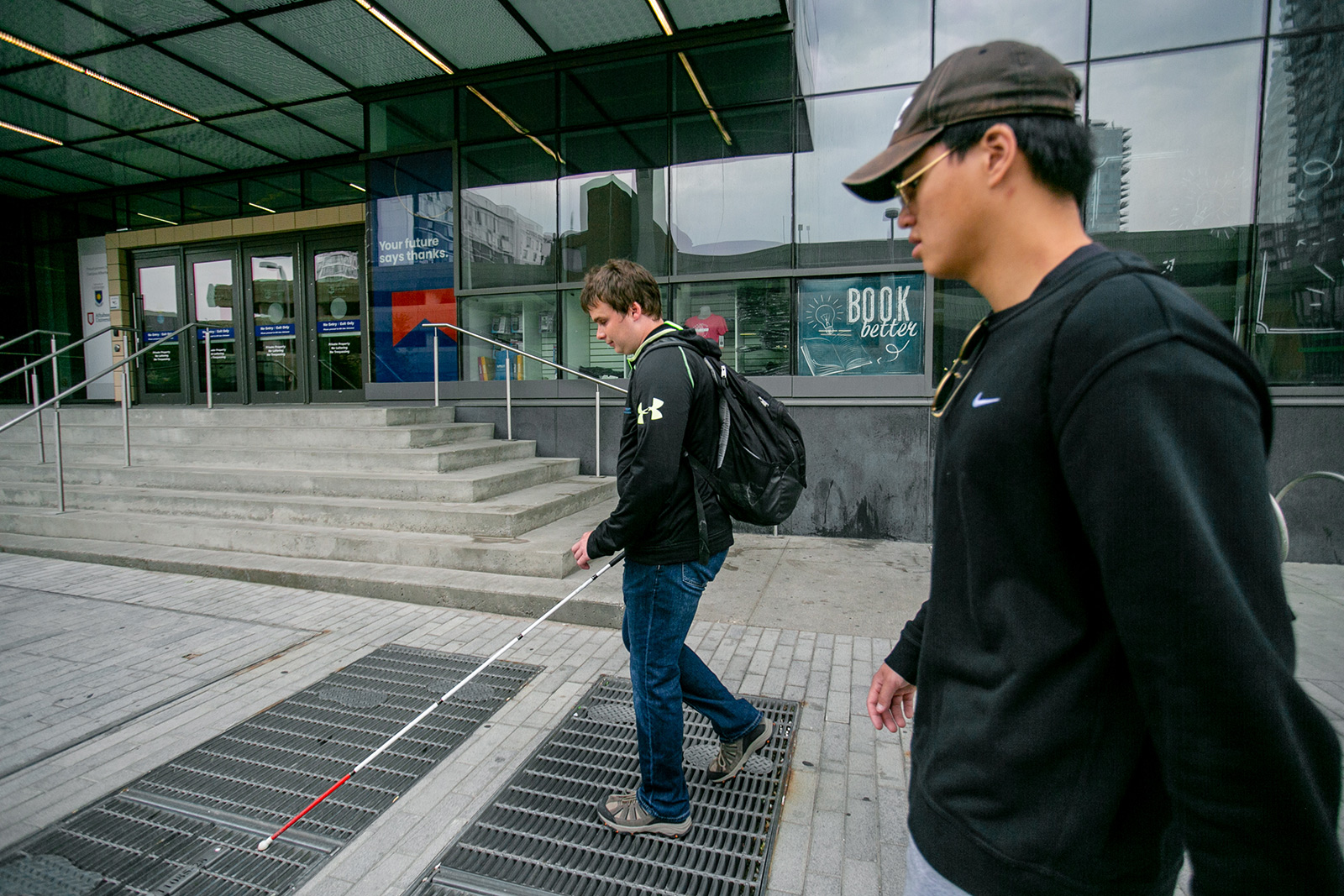 a blind man navigates a city sidewalk with his white cane alongside his non-disabled colleague. In the background is a building with stairs to enter.