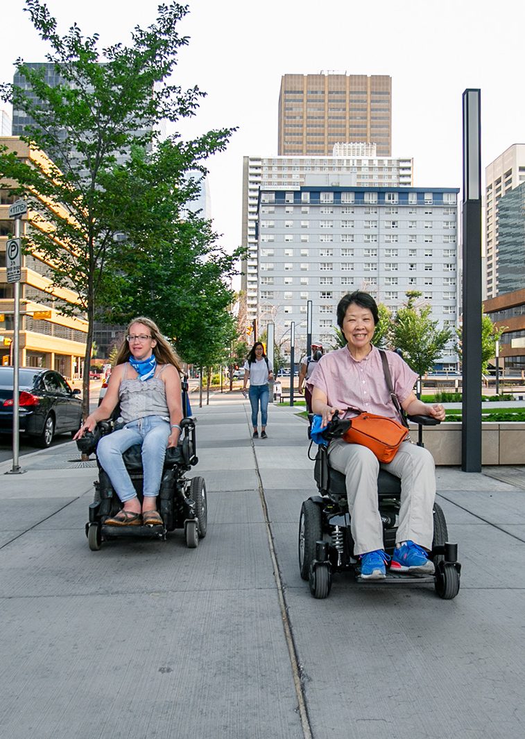 two power wheelchair users drive toward the camera smiling on a city sidewalk