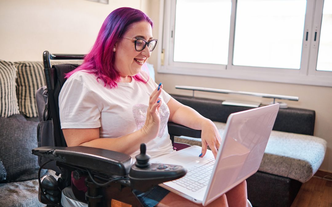 adult woman with pink hair and glasses uses her laptop on her lap while sitting in her wheelchair at home.