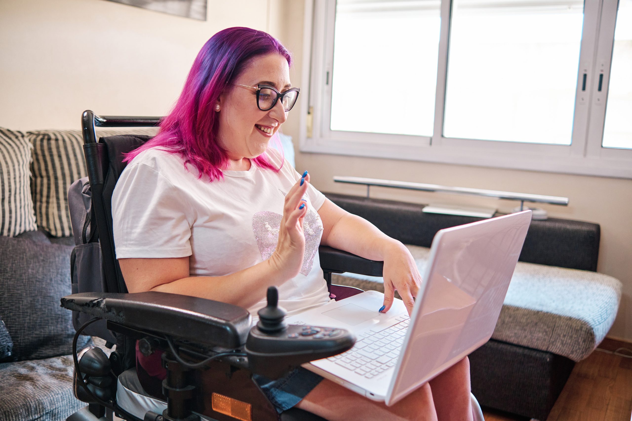 Une femme adulte aux cheveux roses et aux lunettes utilise son ordinateur portable sur ses genoux tout en étant assise dans son fauteuil roulant à la maison.